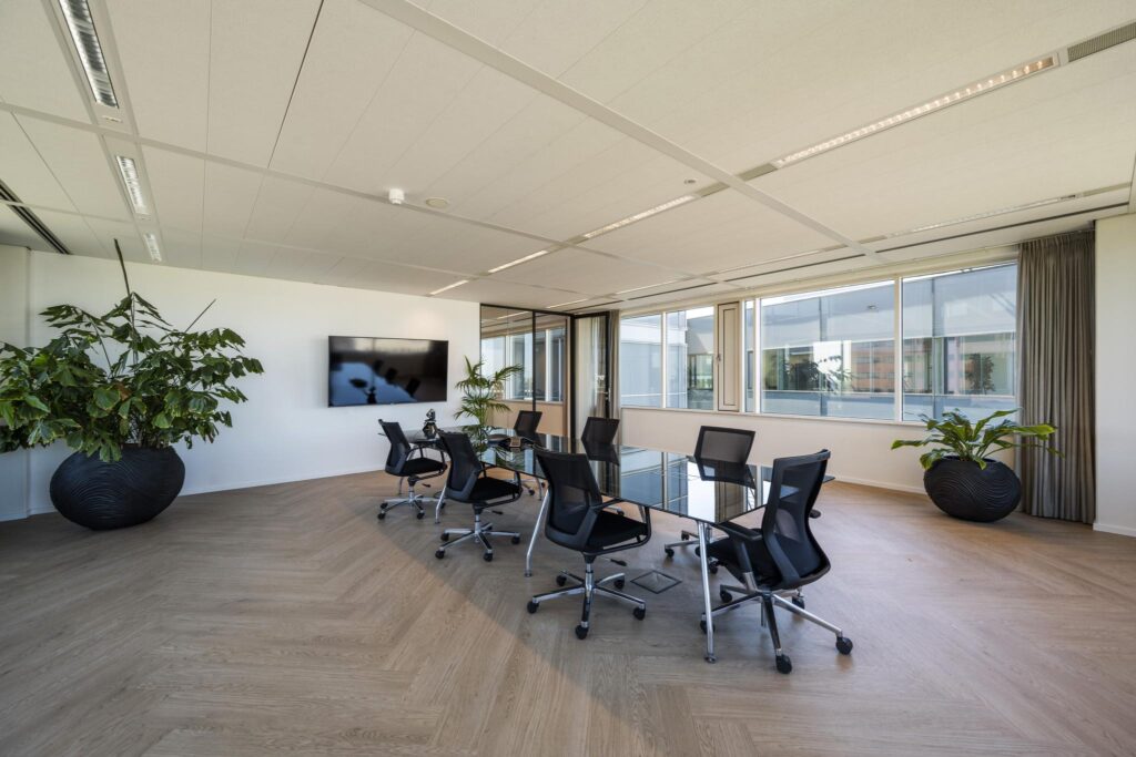 Moderne vergaderruimte aan de Thomas R. Malthusstraat met glazen tafel, ergonomische stoelen, grote planten en een flatscreen aan de muur.